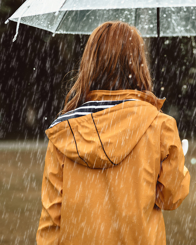 Jente i gul regnjakke som står i regnet under en paraply. Foto